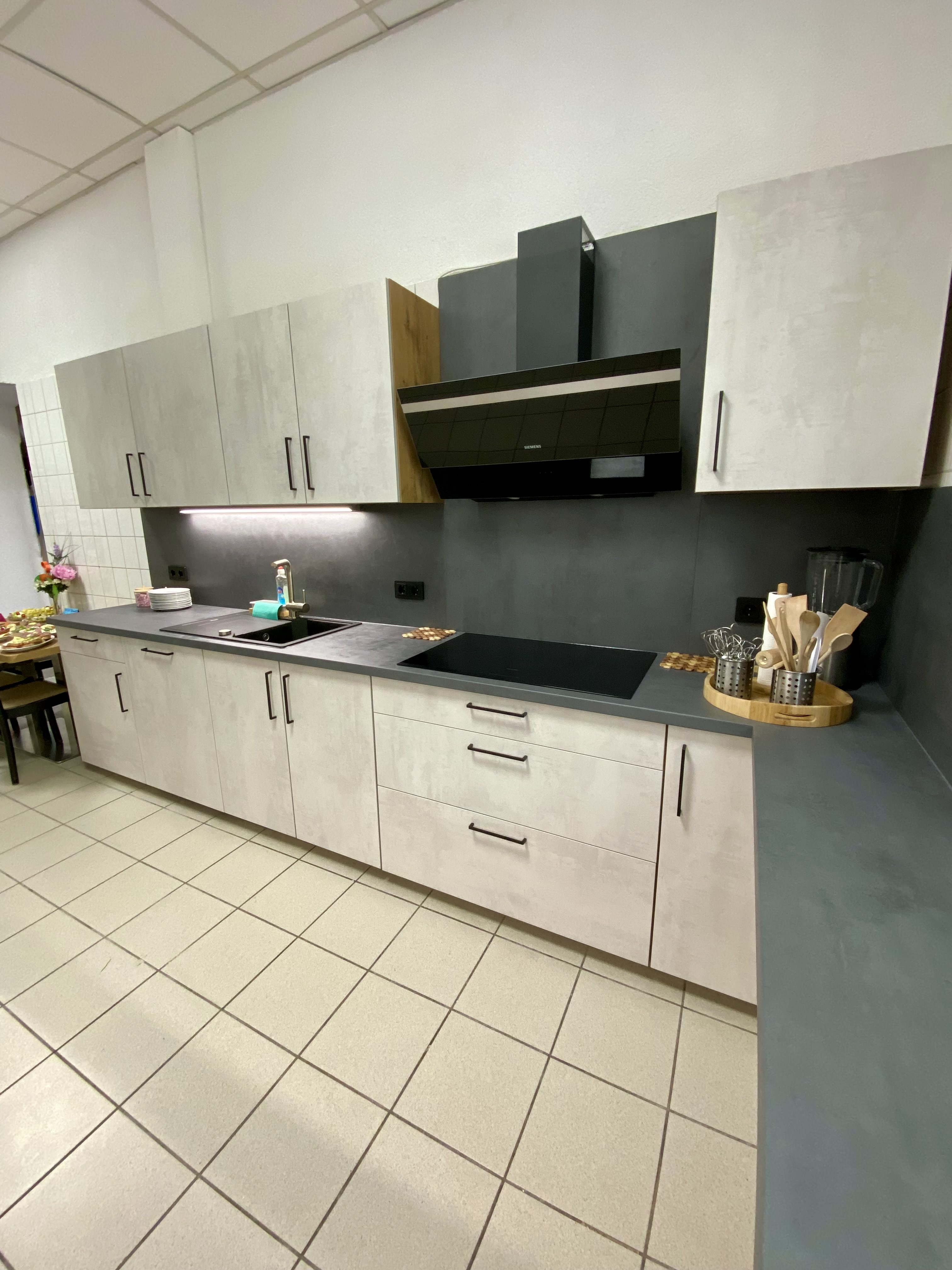 Eine moderne Küchenzeile mit grauen Schränken und einer schwarzen Arbeitsplatte, ausgestattet mit einer Spüle, einigen Küchenutensilien und einer Dunstabzugshaube. Der Raum hat geflieste Böden und ist in neutralen Farbtönen gehalten.