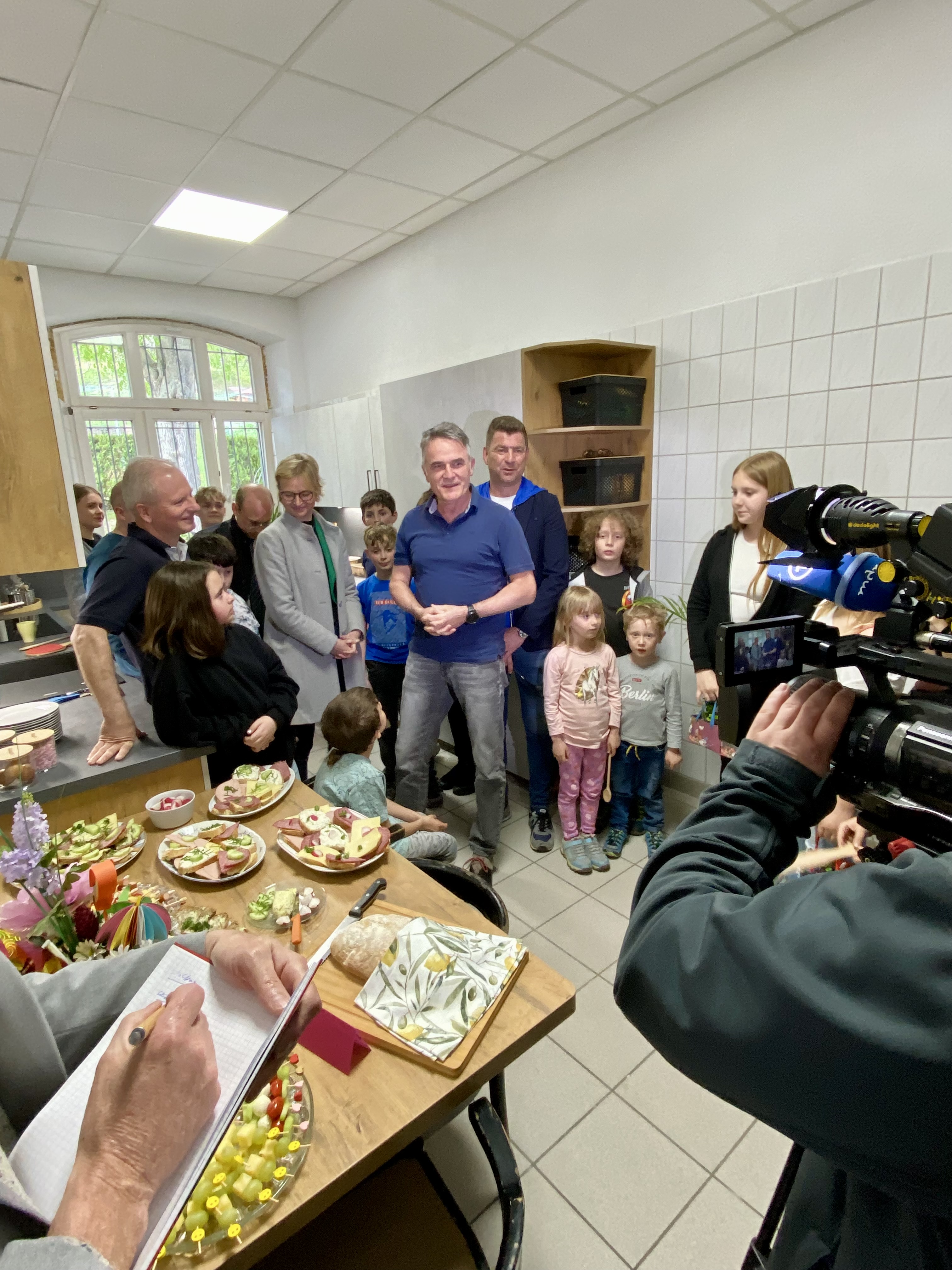 Ein lebhaftes Foto aus einer Küche, wo eine Gruppe Erwachsener und Kinder um einen Tisch mit Häppchen und einer Person, die ein Mikrofon hält, versammelt ist. Ein Kamerateam filmt die Szene. Die Stimmung scheint fröhlich und ausgelassen zu sein.