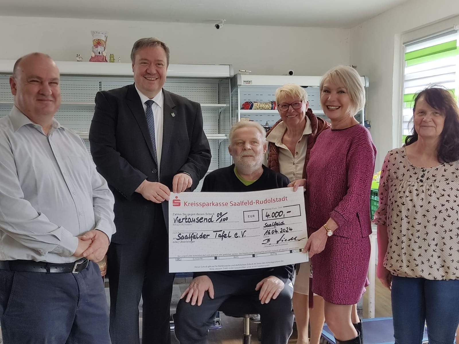 Ein Gruppenfoto von sechs Personen in einem Innenraum, die lächelnd einen großen symbolischen Scheck der "Kreissparkasse Saalfeld-Rudolstadt" über 4.000 Euro präsentieren. Eine Person sitzt, die anderen stehen. Sie scheinen zufrieden und stolz über die Spende.